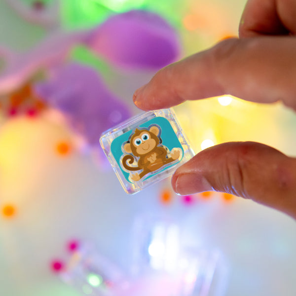 Main tenant un cube lumineux Zany Zoo de Glo Pals illustré d’un singe sur fond turquoise, au-dessus d’un bac de jeu sensoriel.