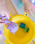 Mains manipulant un outil violet au-dessus d’une passoire jaune en forme de crabe, dans un bac d’eau avec cubes lumineux Glo Pals.