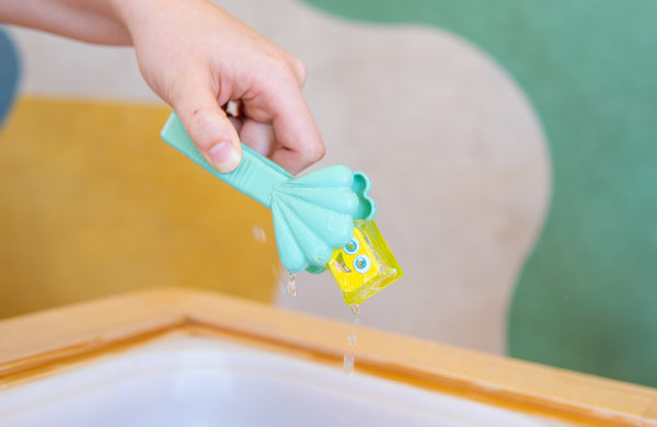 Main tenant une pince vert menthe en forme de coquillage pour saisir un cube jaune Glo Pals au-dessus d’un bac d’eau.